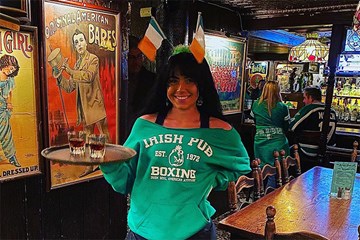 Waitress at the Irish Pub holds two shot glasses on a tray with Irish flag head band and green boxing sweatshirt.