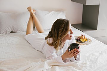 Woman lounging in comfort on hotel bed drinking warm beverage with plate of fresh fruit nearby.