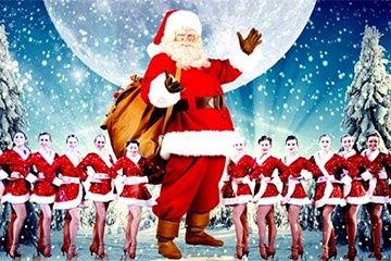 Santa stands with sack of gifts with showgirls dressed in Santa outfits behind him.