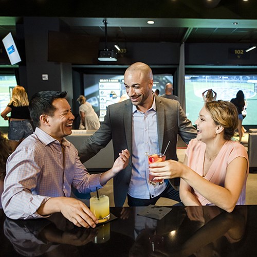 Man and woman seated at bar with drinks in hand while laughing with another man standing.