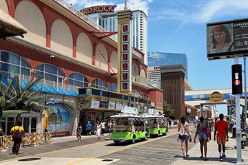People walking on the Atlantic City Boardwalk near Resorts and Hard Rock Casinos. A boardwalk tram is also in view.