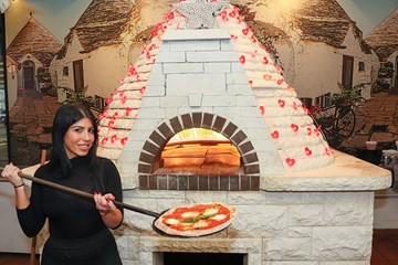 Woman showing a margarita pizza pie with Valentine's Day decor adorning brick oven.